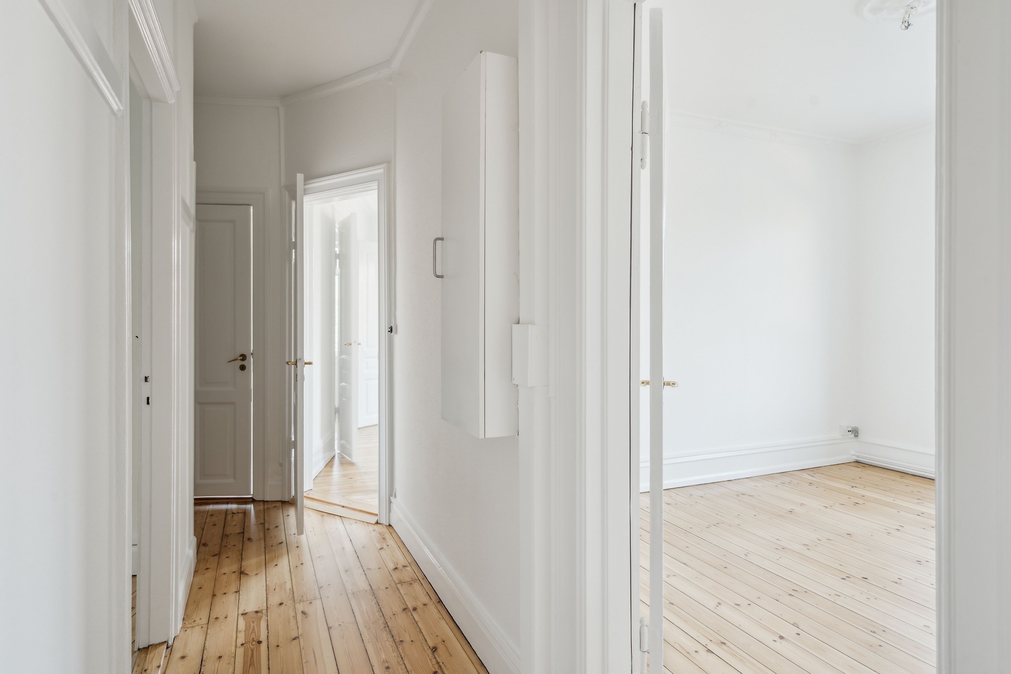 En rummelig korridor med lyse vægge og trægulv, åbne døre og hvide dørkarmer og tavler, trægulvet reflekterer lyset og