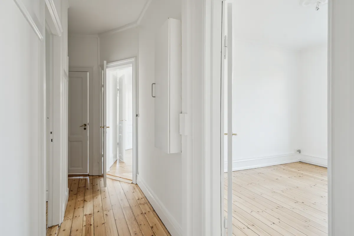 En rummelig korridor med lyse vægge og trægulv, åbne døre og hvide dørkarmer og tavler, trægulvet reflekterer lyset og