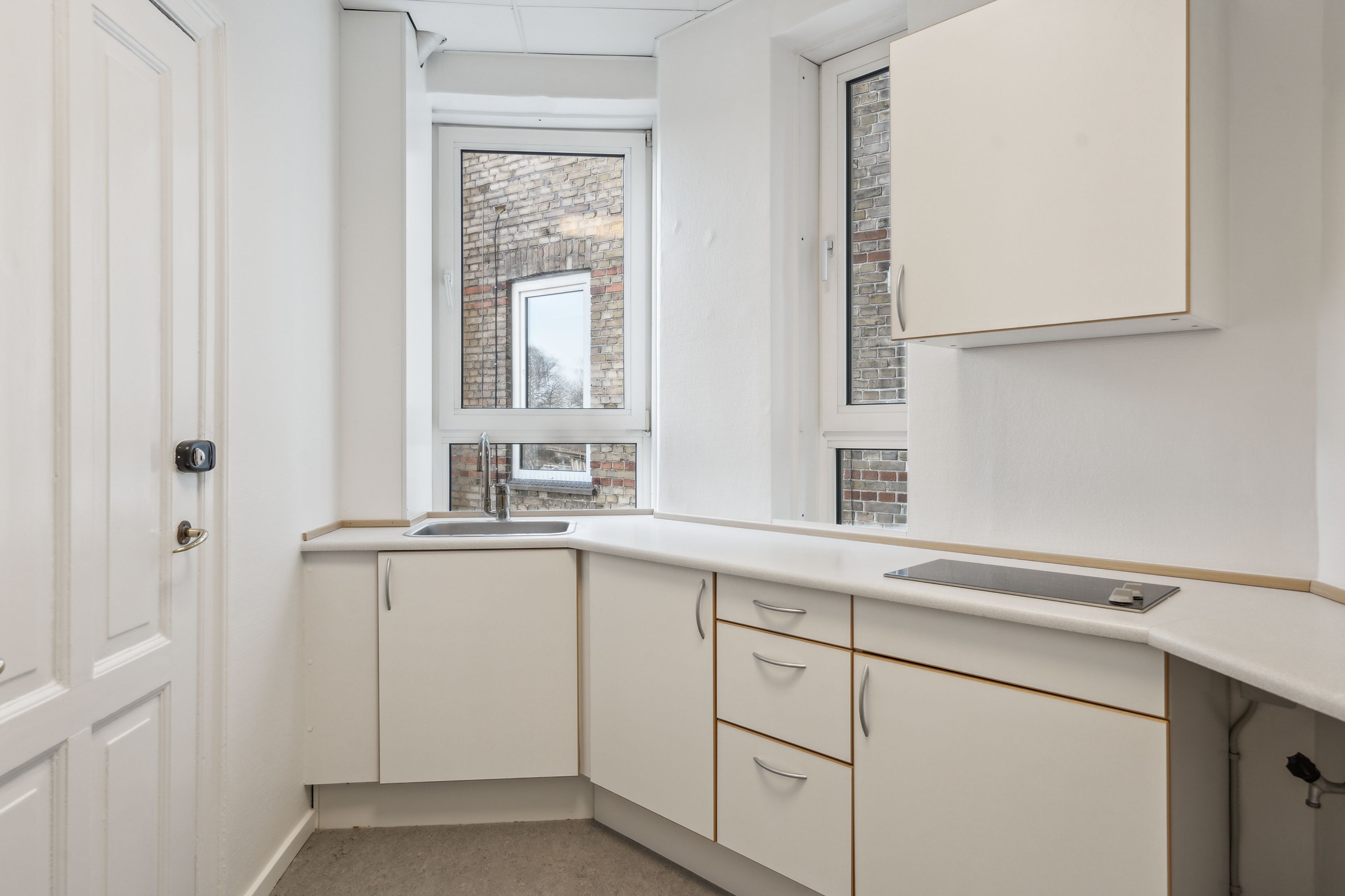 En lille hvid køkken med hvide vægge, hvide skabe, en brændpladeskomfur og et vindue med udsigt til en murstensbygning