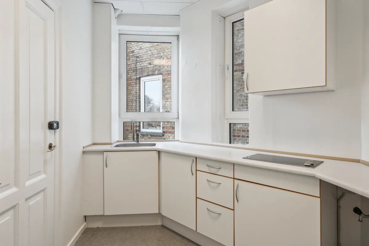 En lille hvid køkken med hvide vægge, hvide skabe, en brændpladeskomfur og et vindue med udsigt til en murstensbygning