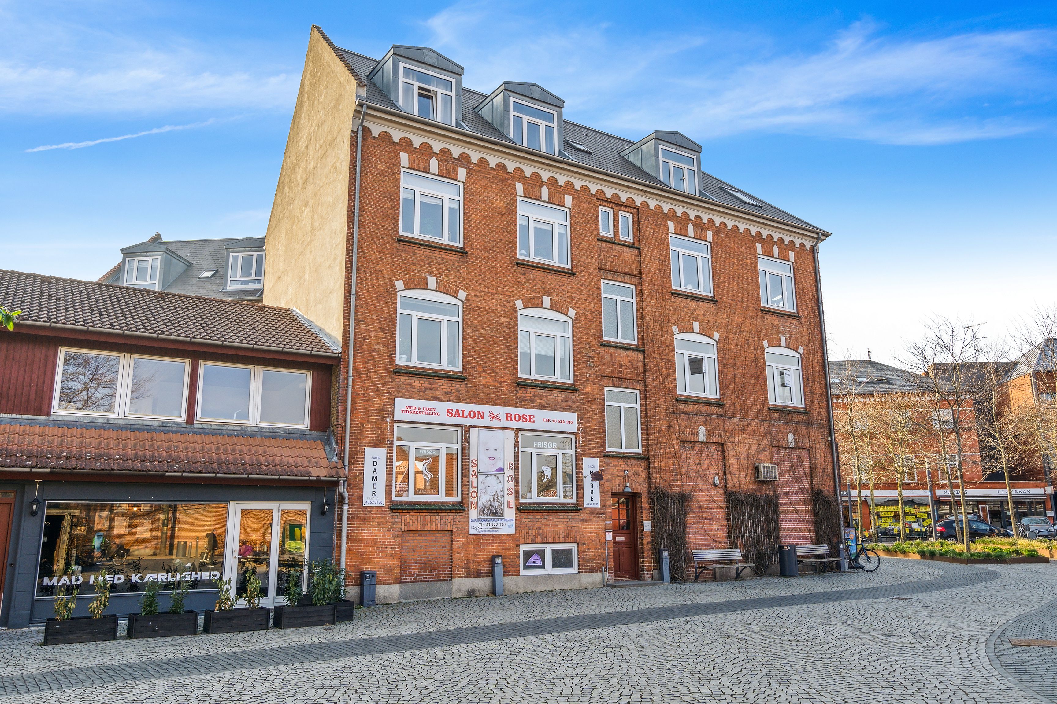 En stor murstensbygning med mange vinduer og en røgfangere, udsmykket med plantepoter og bænke. Der er tekster og skilte på