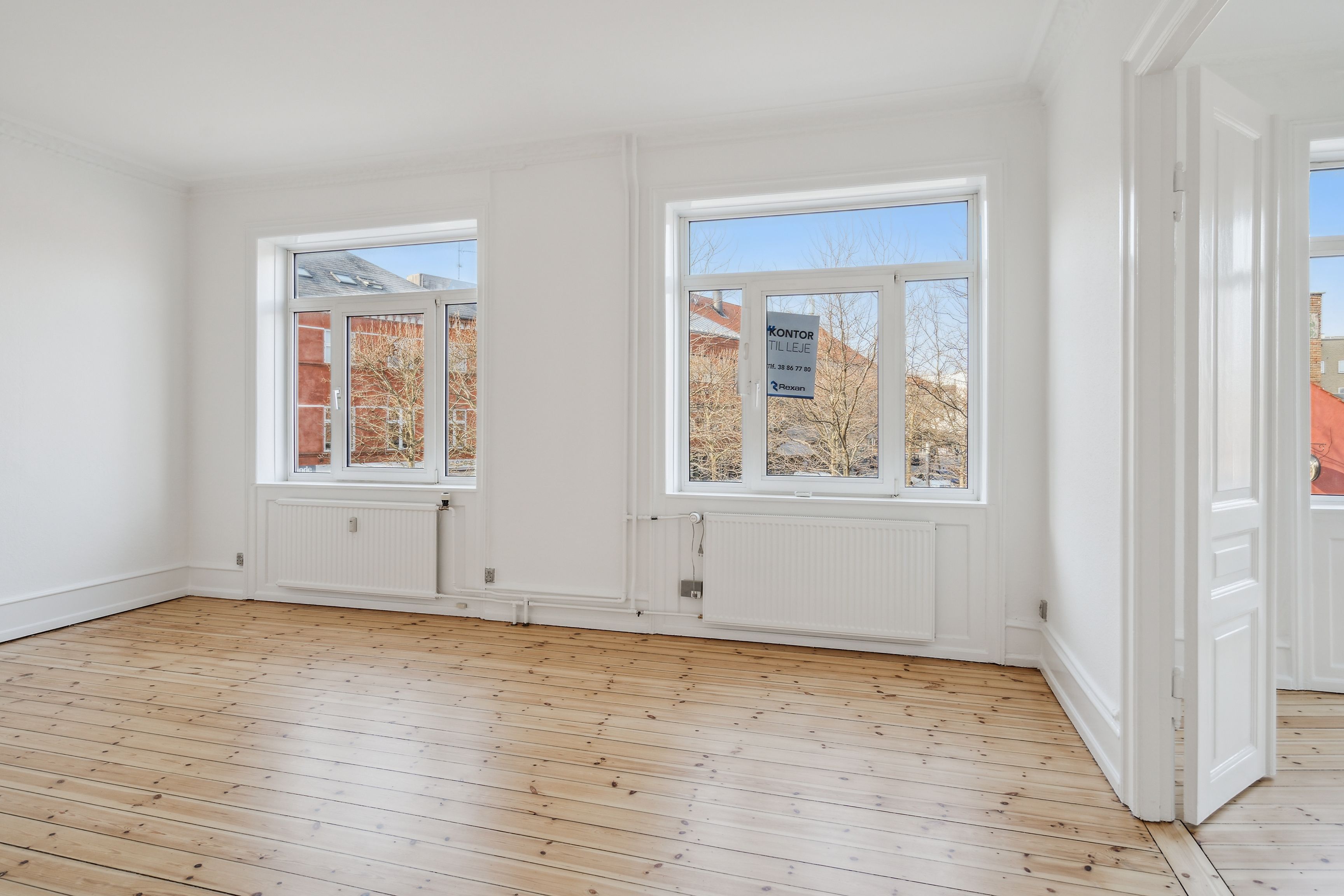 Trægulvede og hvide vægge med tre store vinduer, som giver masser af lys og udsigt til bygninger og træer udenfor