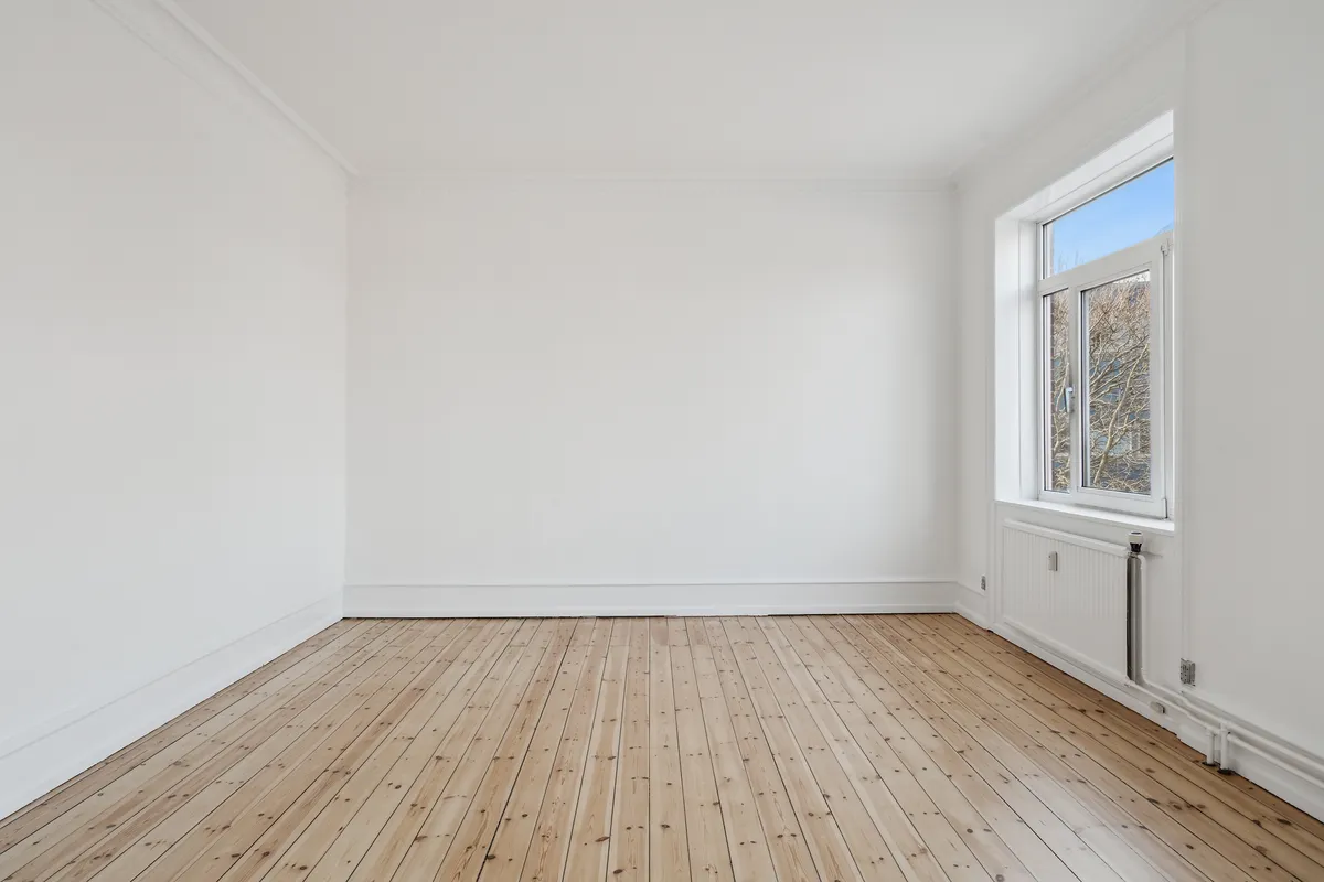 Et tomt, lyse og rent kontor med hvidmalede vægge og trægulv, samt en åben sølvfarvet rude med udsigt til en træstamme