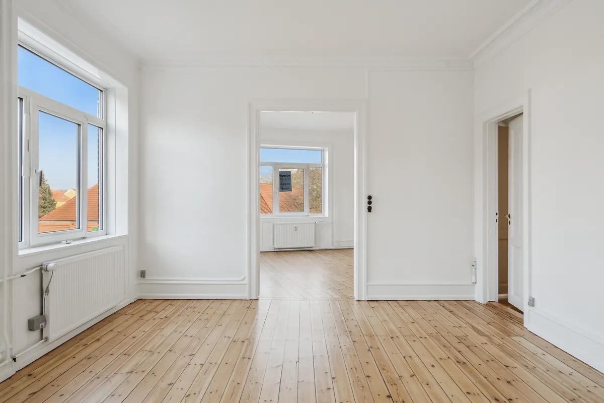 Kontor med hvide vægge, lyse gulvplanker og to store vinduer med udsigt til en grøn skov. To radiatorer er installeret på