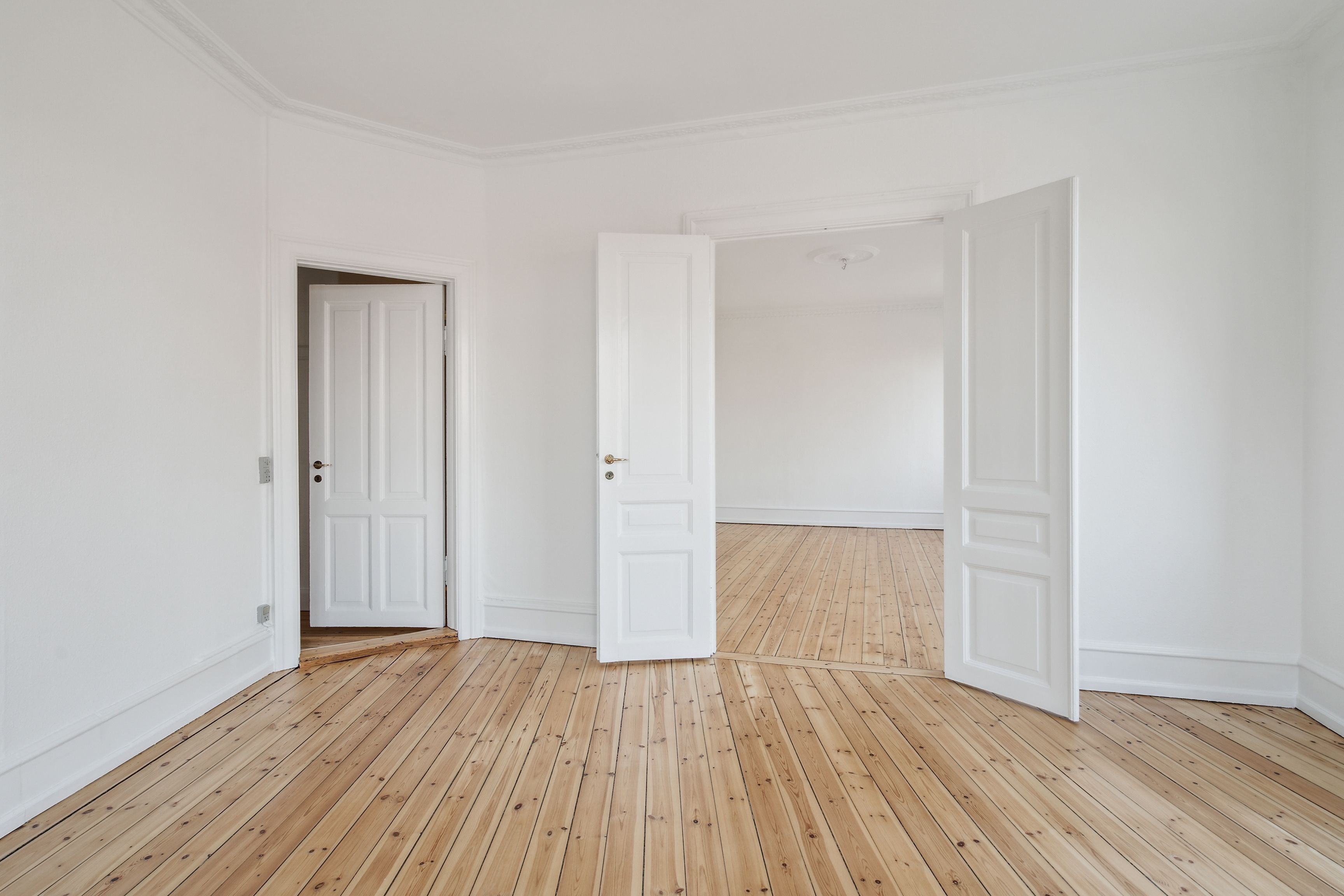 To store hvide døre og en lille dør er åbne i en stor, tom stue med lyse, trægulve og hvide vægge