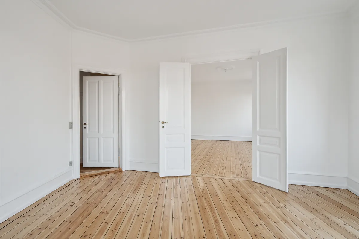 To store hvide døre og en lille dør er åbne i en stor, tom stue med lyse, trægulve og hvide vægge