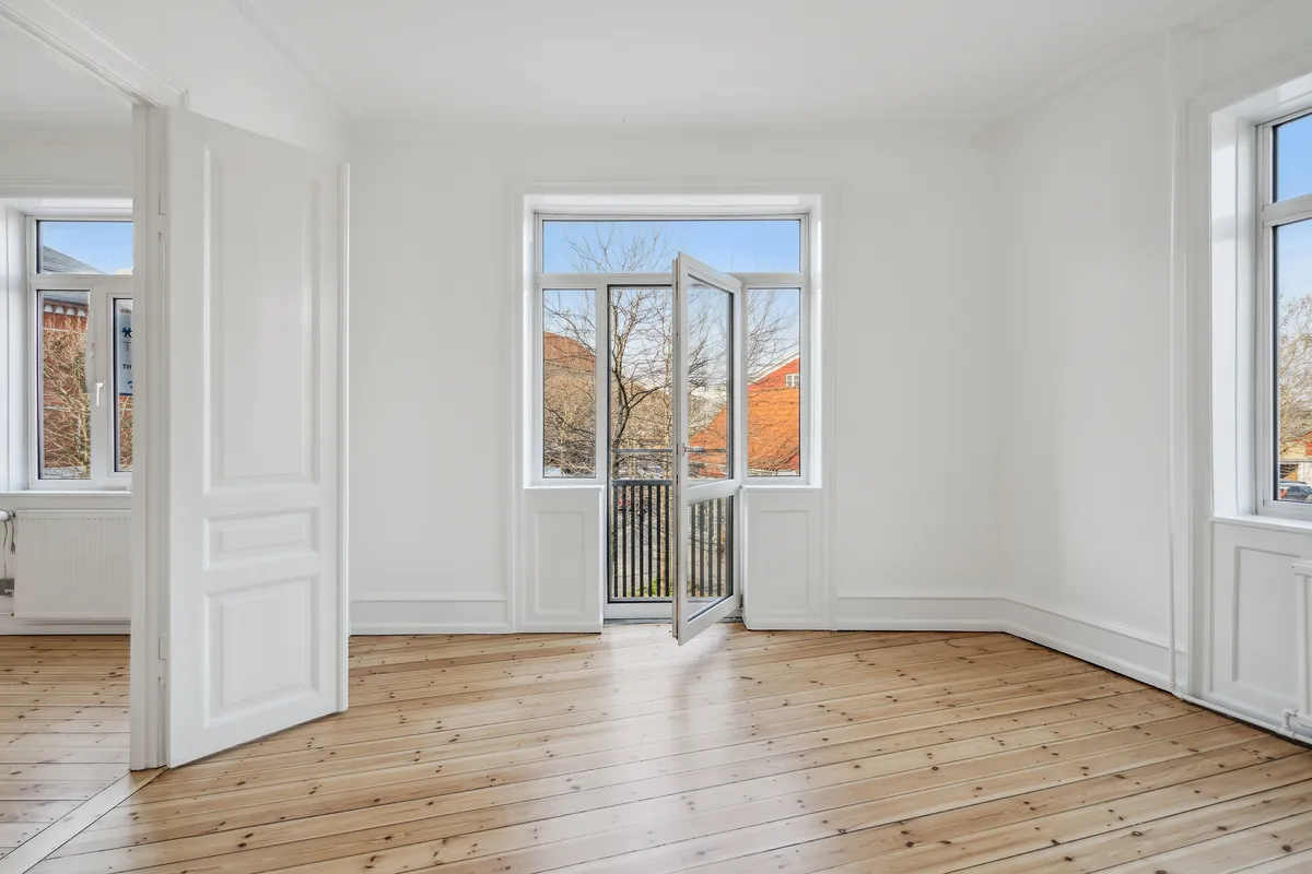Indvendørs rum med hvidmalede vægge, trægulve og åbne døre og vinduer. To vinduer med glas og en balkon med metalgitter.