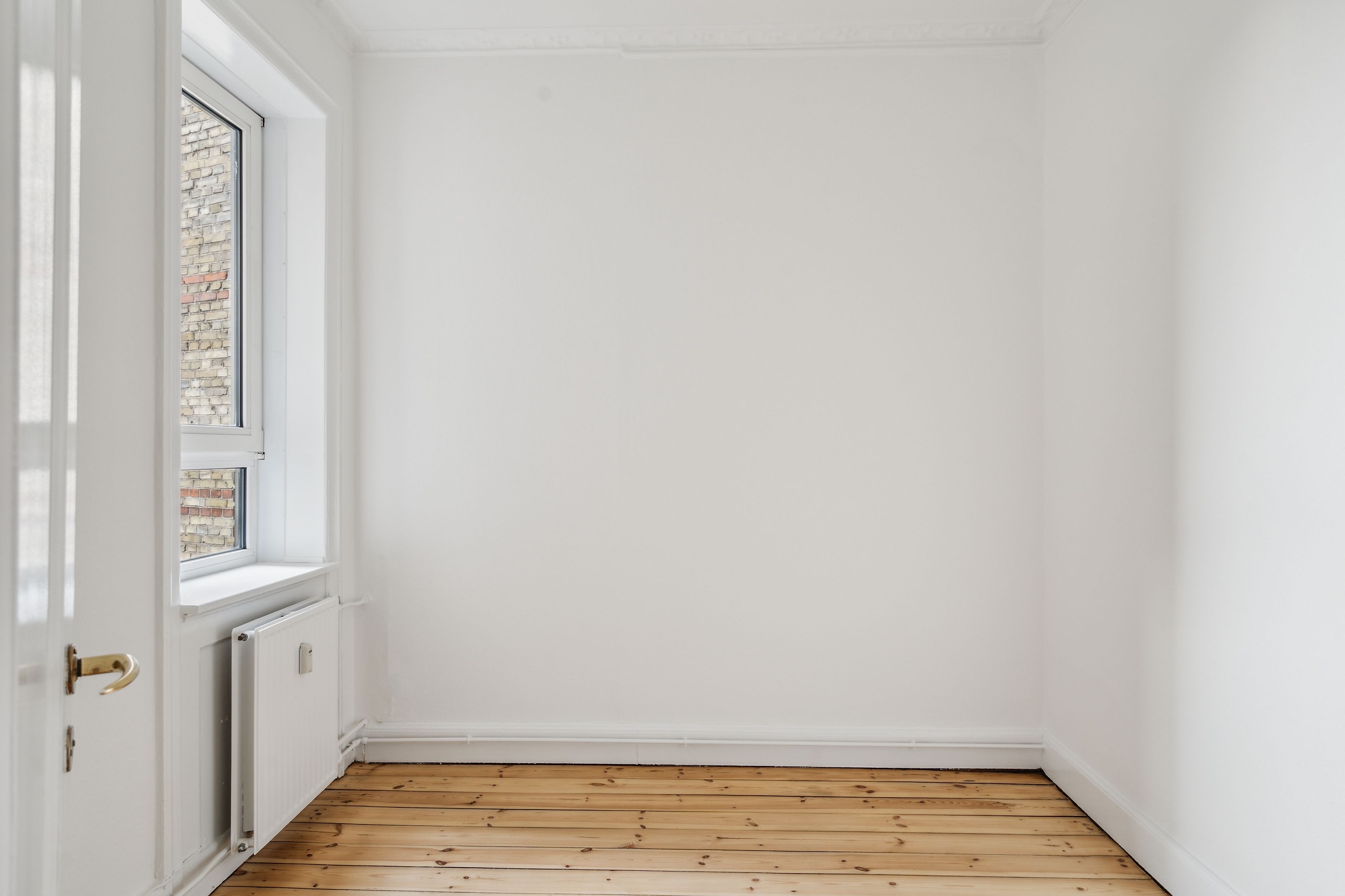 En stor, tom stue med hvide vægge og lyse trægulve samt en radiator og et vindue med udsigt til en murstensbygning