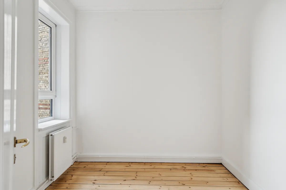 En stor, tom stue med hvide vægge og lyse trægulve samt en radiator og et vindue med udsigt til en murstensbygning