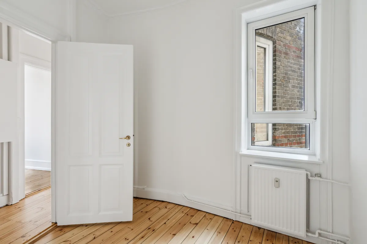 Et rum med hvide vægge og trægulv, der er åbnet mod et andet rum, hvor der er et hvidt vindue og en radiator