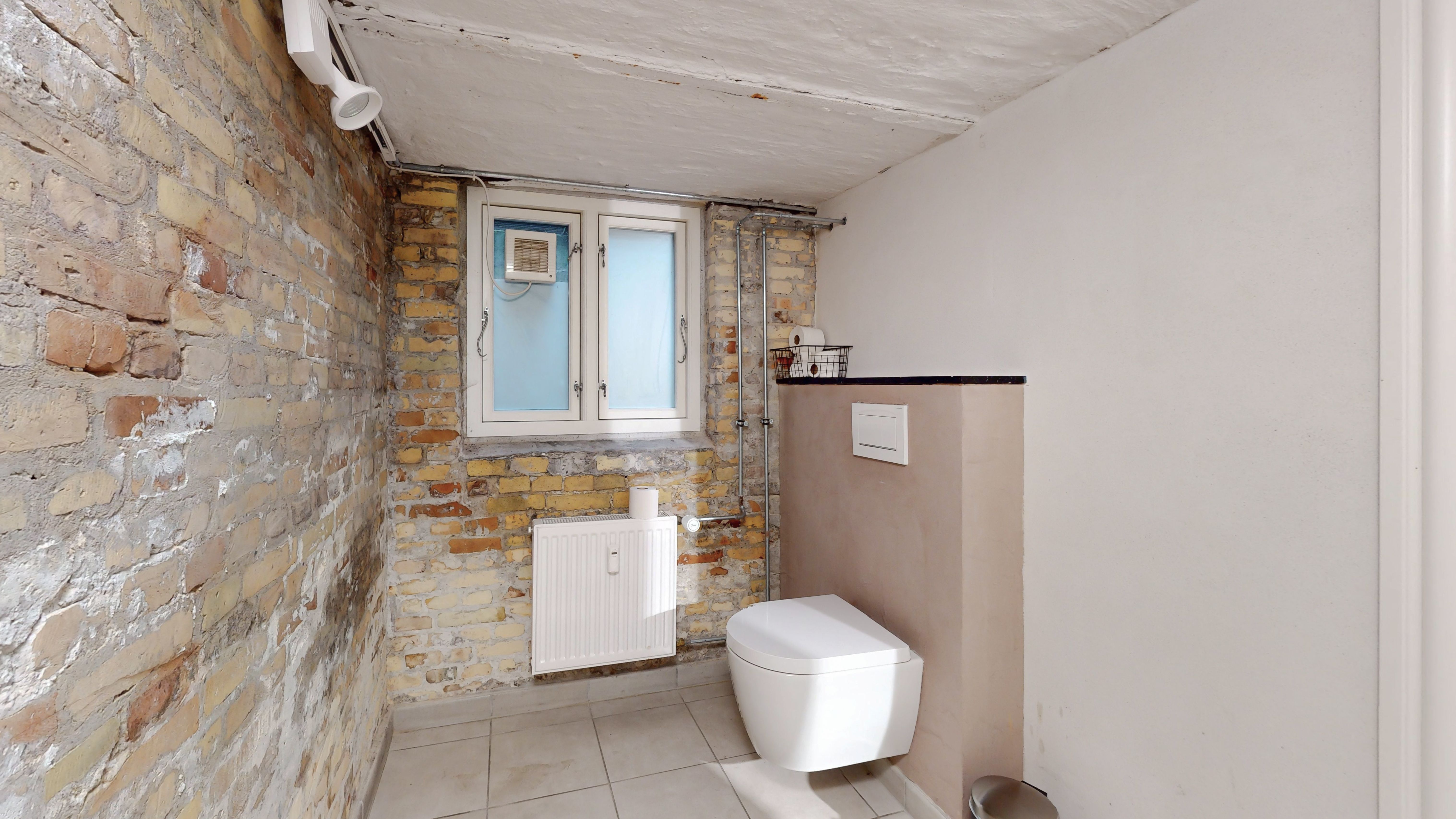 En toilet med lyse, moderne interiør, hvide keramiske toilet, og en lysegrå væg. En radiator er monteret på væggen, og der