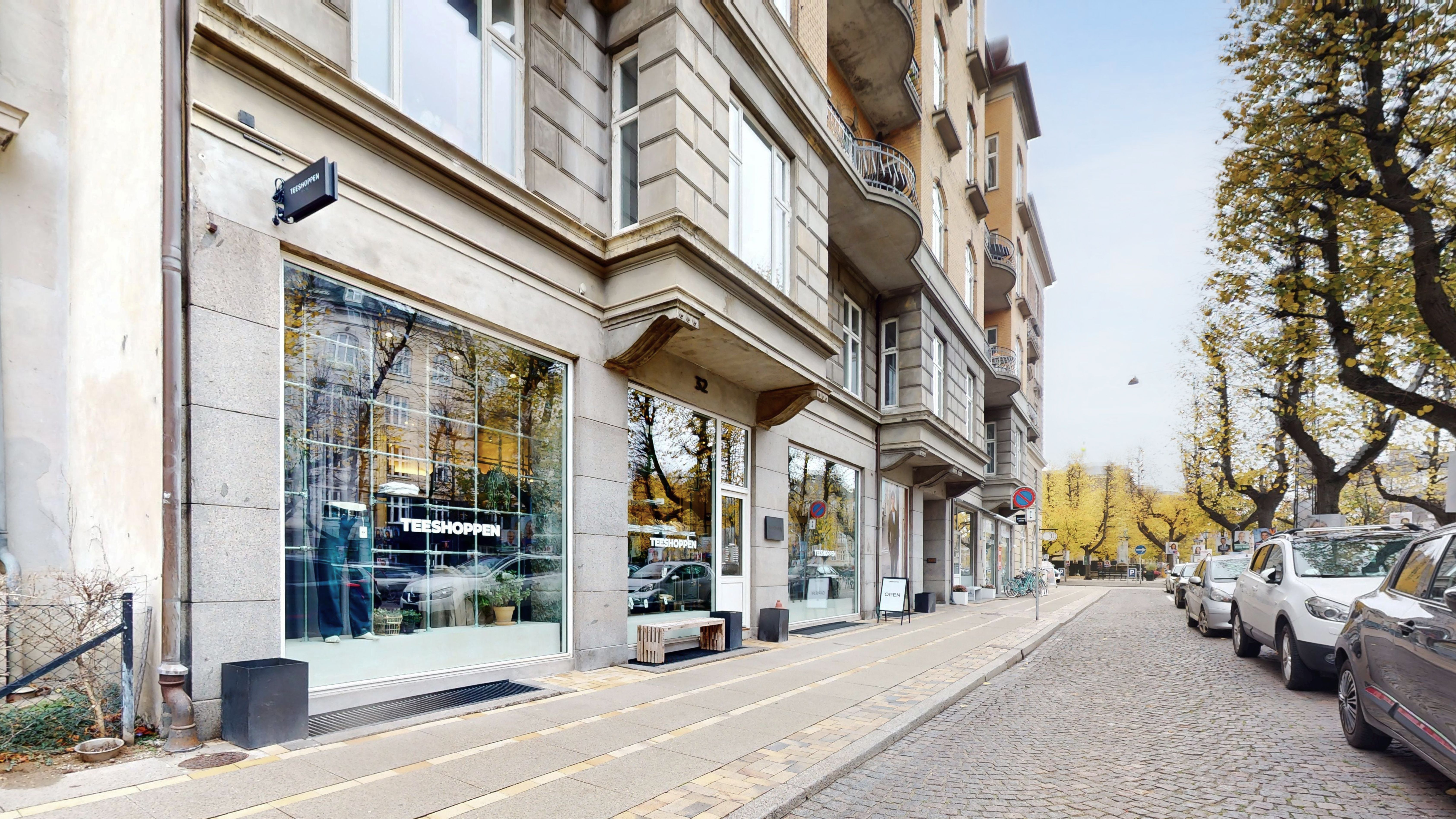 Frederiksberg Allé med butikker, træer og parkerede biler på en kollektiv buskørselsvej og et snedækket grøntområde