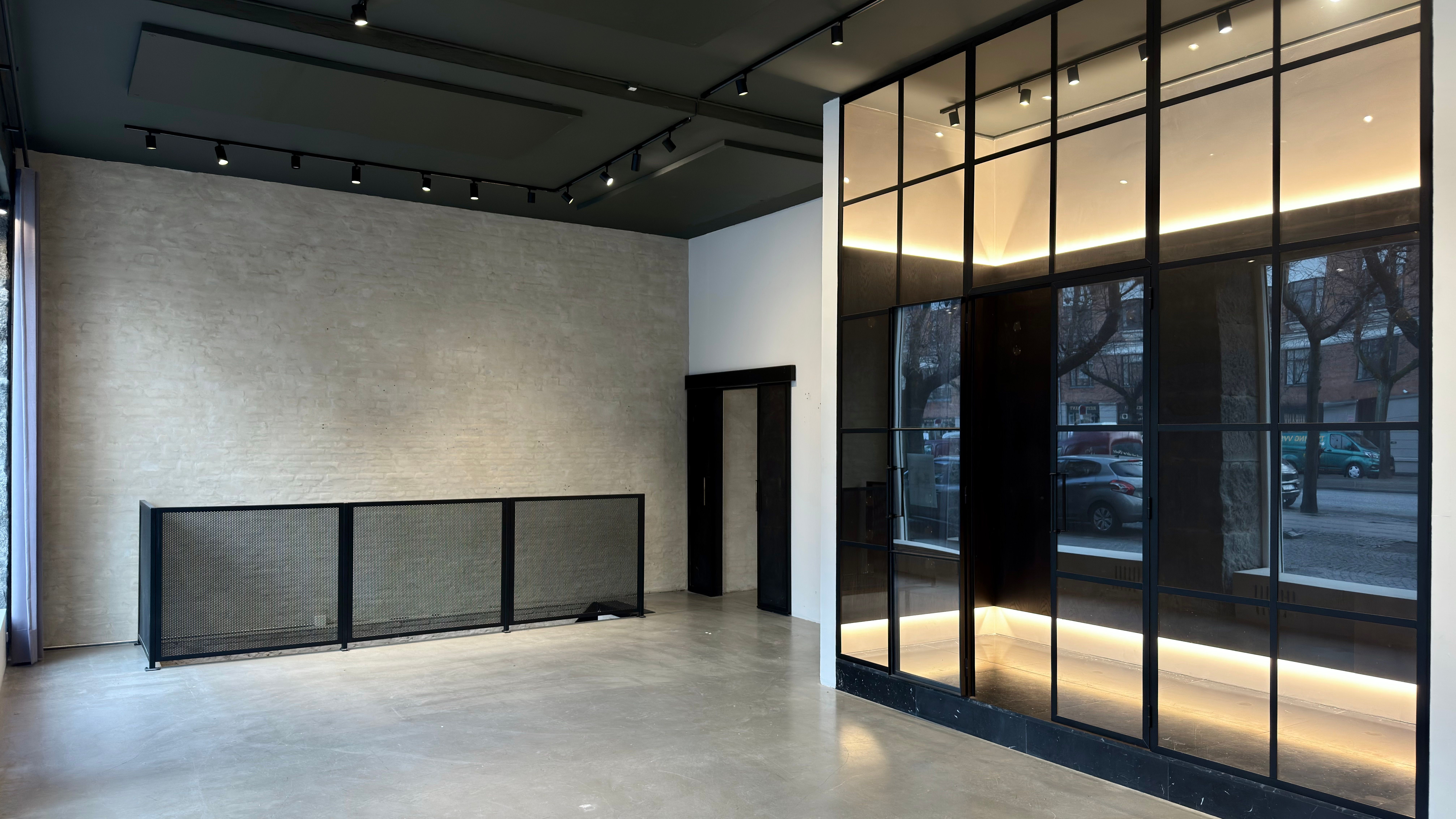 Store glasruder og mørke metalprofiler giver en moderne og elegant indgangszone til et kontorlokal med betongulv og lyse accenter på væggen