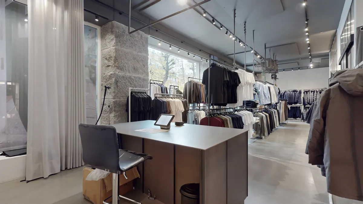 En butik med klæde og tøj på hylder og søjler. En tablet er på en bordplade, og en stol står til siden. Hvidt og gråt er