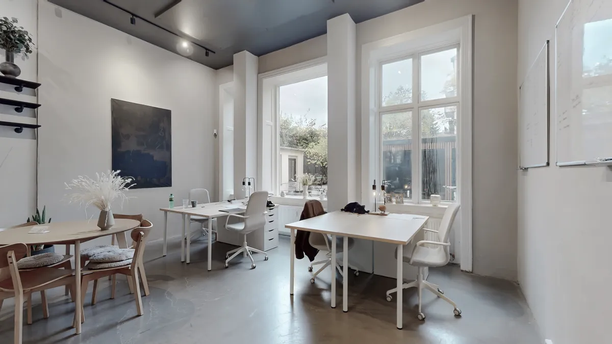 Hvidt og rent kontor med to skriveborde og stole samt et større skrivebord med to stole og plante på skrivebordet