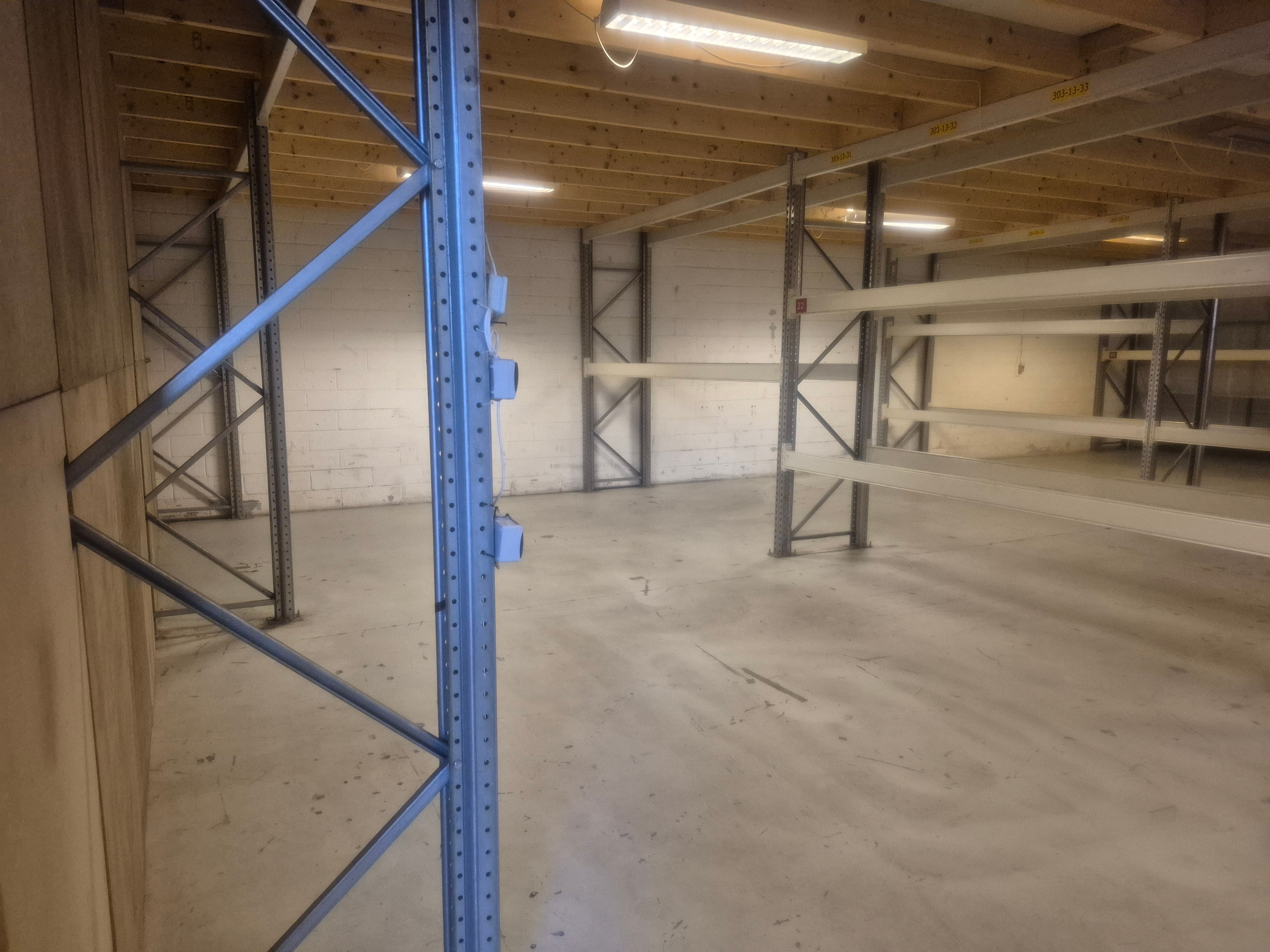 Et stort og tomt lager med stålreoler og hvide vægge og lyse gulve med et metalrør og elektriske ledninger på væggen