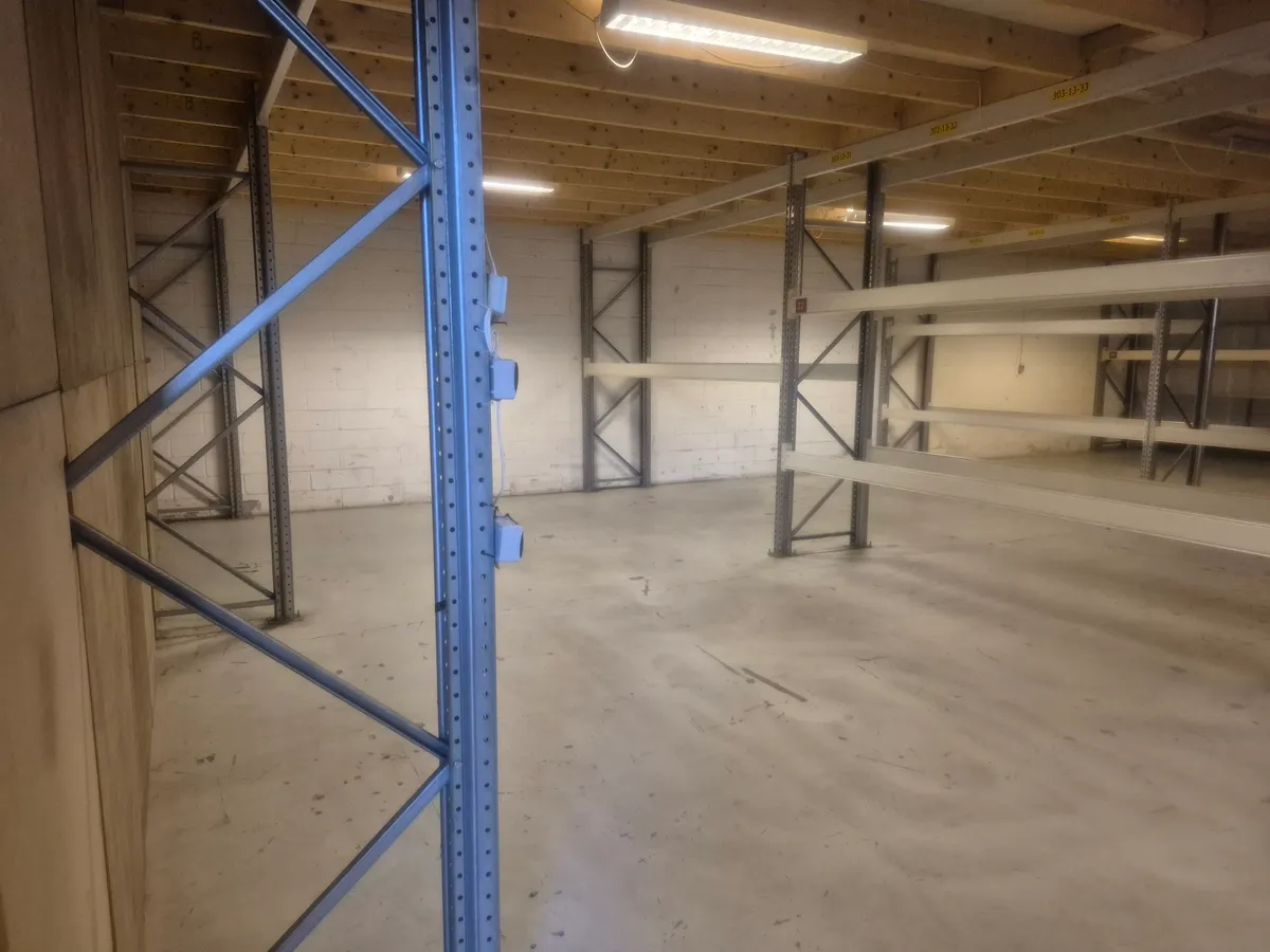 Et stort og tomt lager med stålreoler og hvide vægge og lyse gulve med et metalrør og elektriske ledninger på væggen