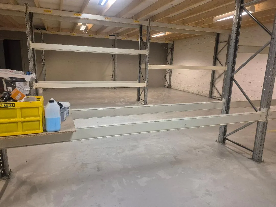 Lager med metalliske regler, hvor gulvet er lige. Der er lyse og to palleløfter. En metro-kasse og en plastflaske med væske står på loftet.