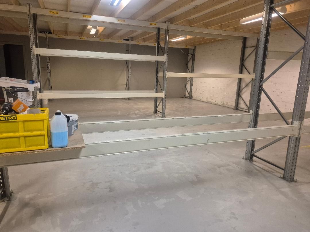Lager med høje metalreoler og gulv. Forskellige lagervarer og et håndværktøjskabs er placeret på paller og i kasser.