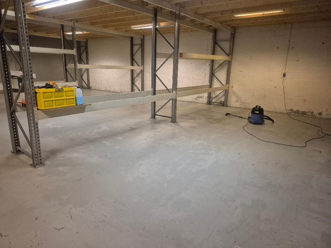 Lager med metalreoler og et gult plastkasse. Væggene er hvide og der er gul linje på gulvet. Et støvsuger er placeret på gulvet.