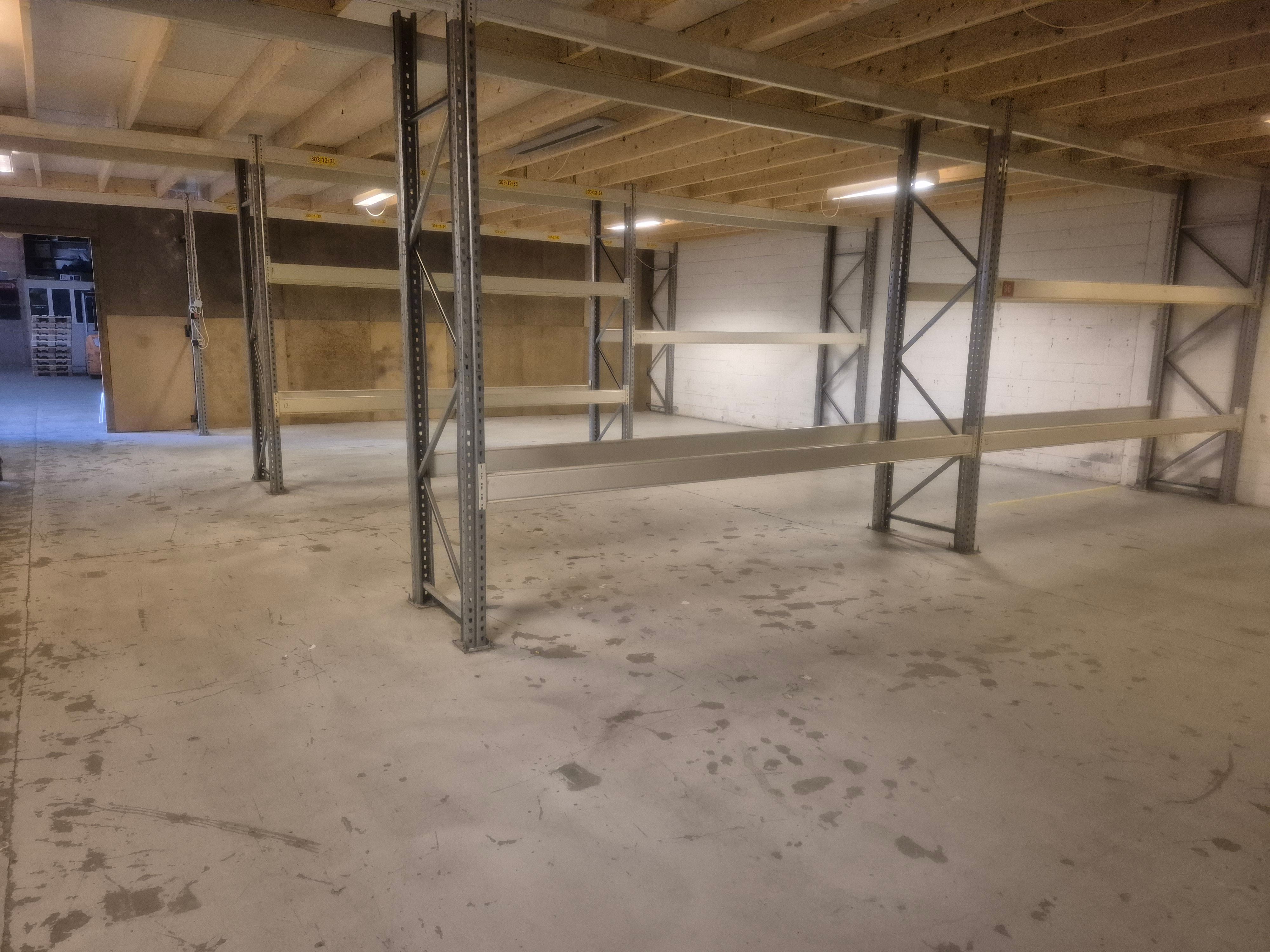 Lager med flere tomme hylder og højt loft, to lyse på loftet og en dør i baggrunden