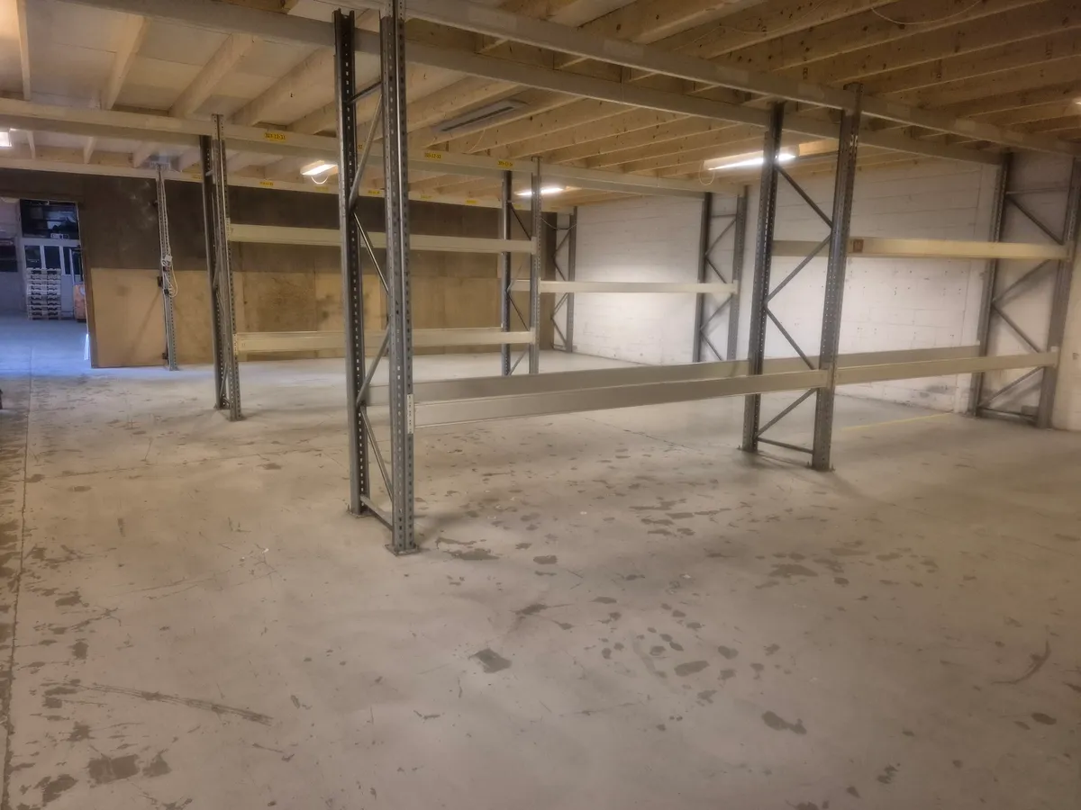 Lager med flere tomme hylder og højt loft, to lyse på loftet og en dør i baggrunden