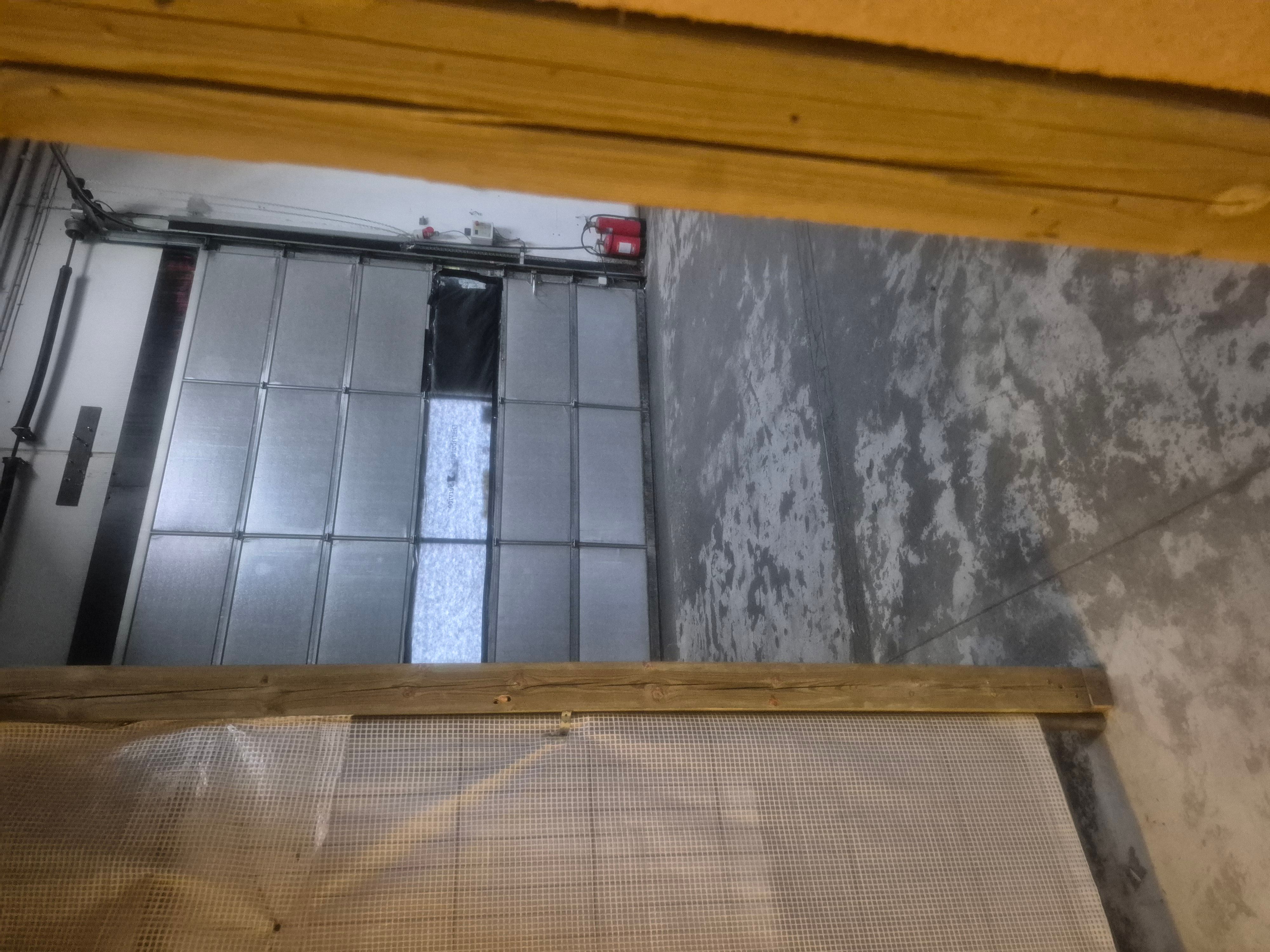 Glasdør i garage med rødt brandslukker og skraldkasse samt et stykke netværk uden for døren