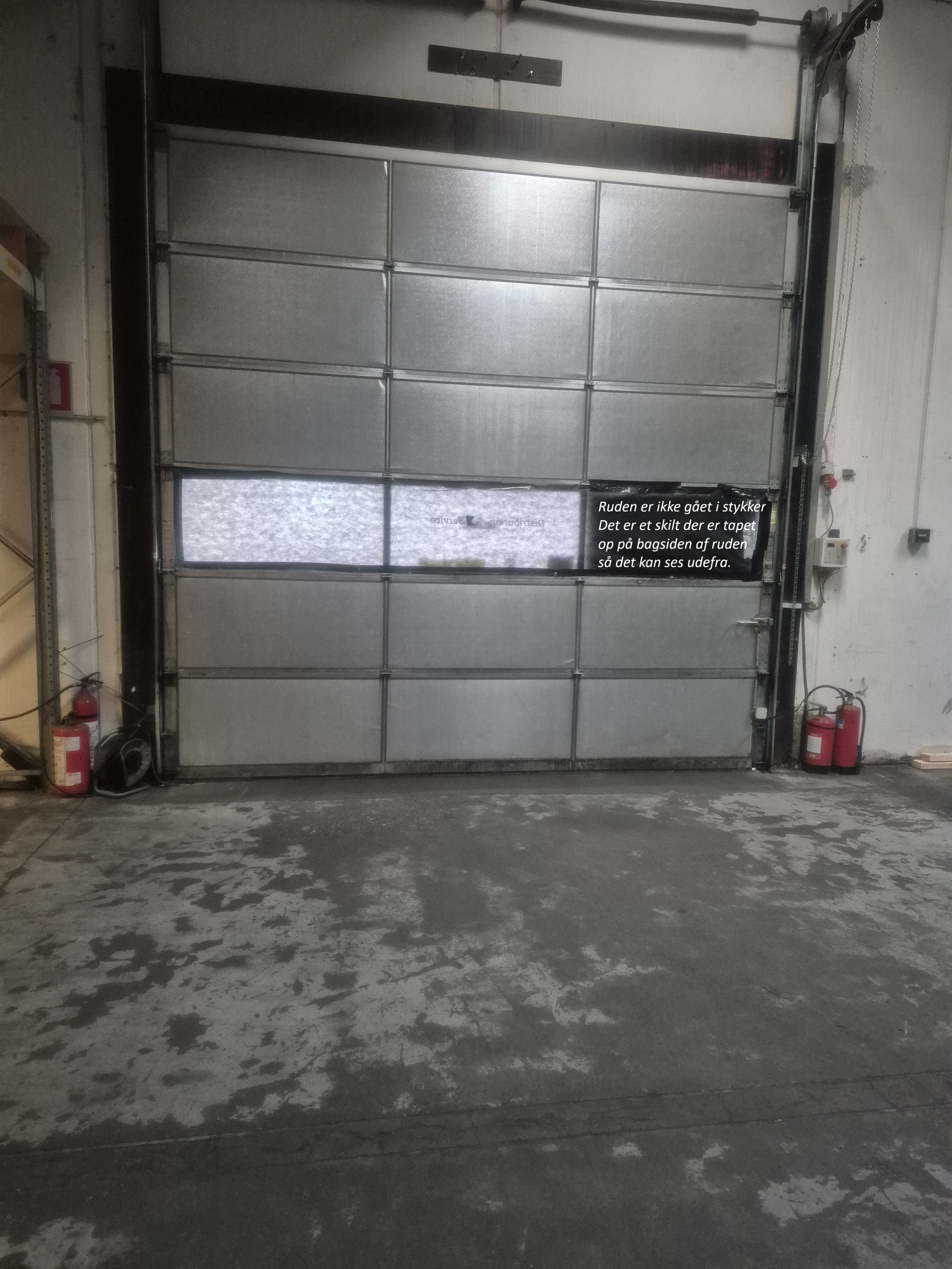 Et stort, hvidt garageloft med automatisk garagedør og to ildslukker. Der er en tekst, der står "Ruden er ikke gået i stykker" på døren