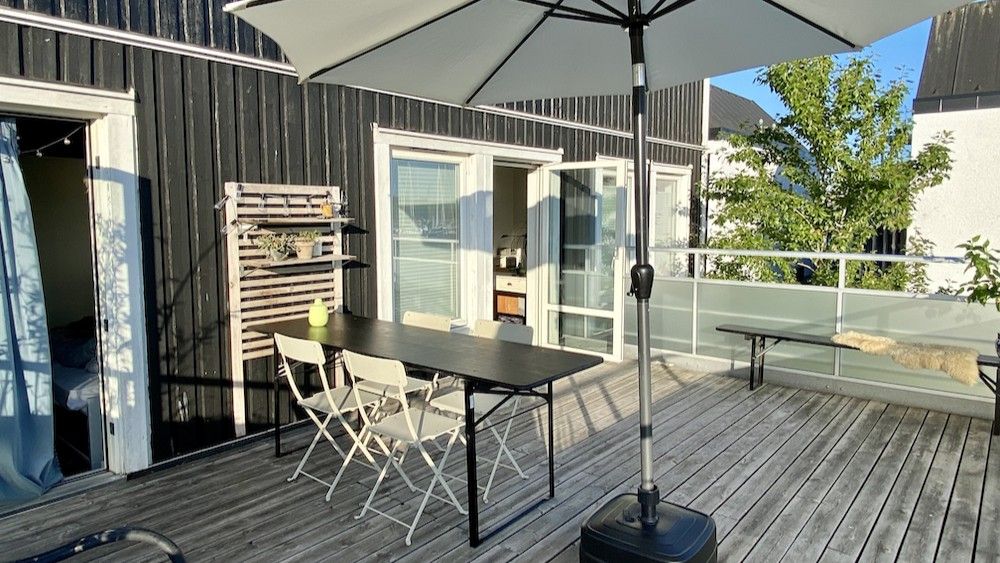 En stor parasol er ophængt over et bord med fire stole på en træterrasse og en parkeringsplads