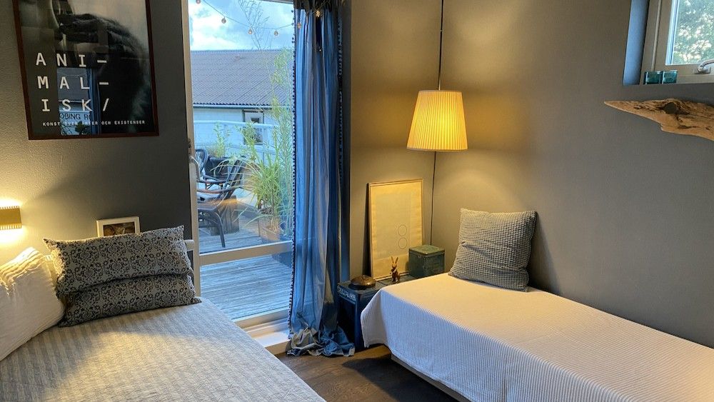 To soveværelser med enkeltsenge og vinduer. Der er lysekroner og vægdekorationer. En sofa og bord med en vase. En