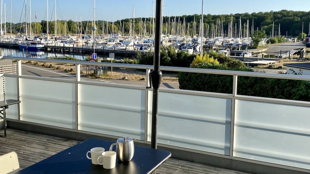 Terrasse med udsigt over havn med mange både og maste. På bordet er der to krus og en kaffekar med mælk.
