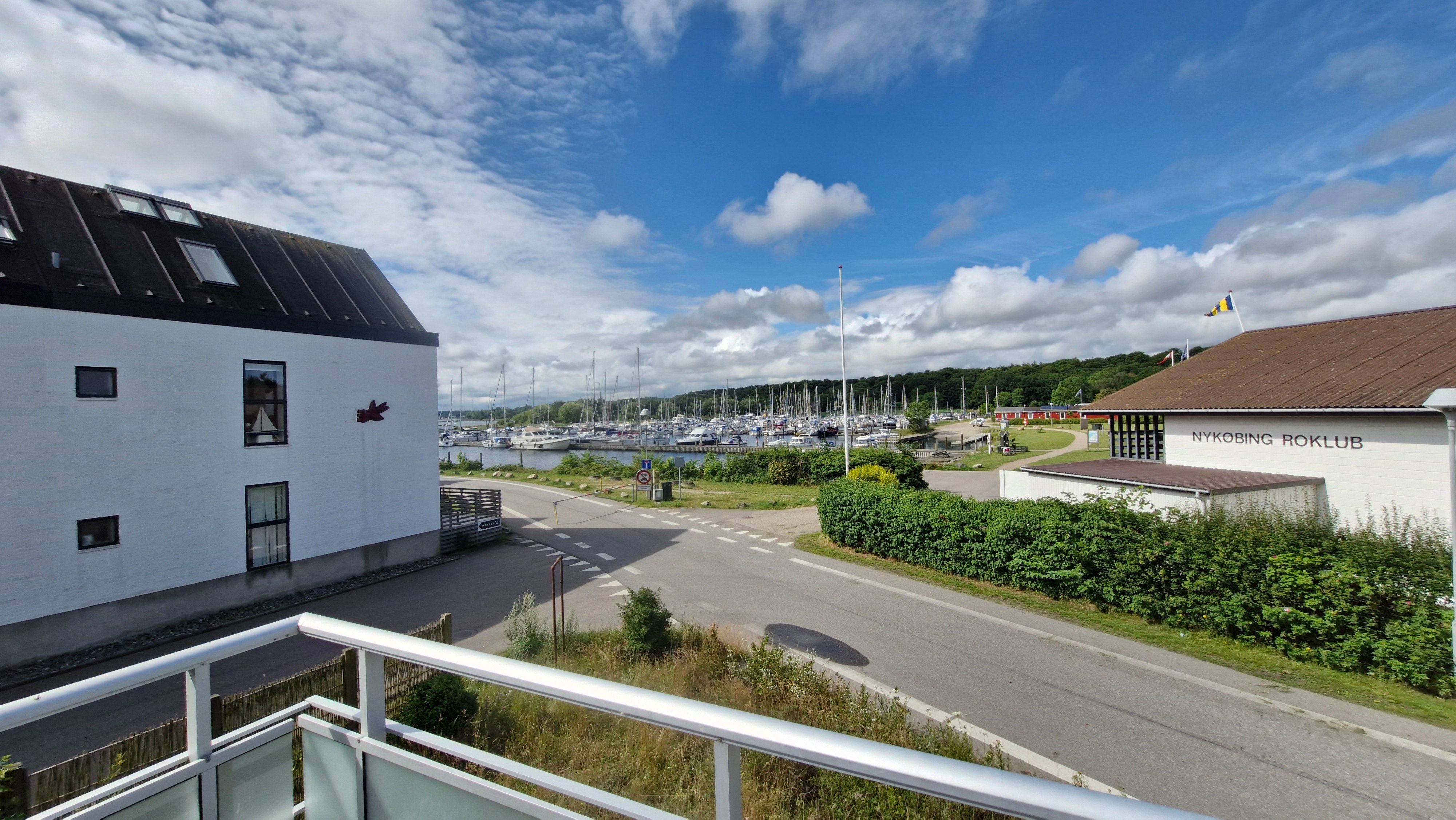 Et luftigt hav med adskillige både og Nykøbing Roklub på højre side af en asfaltvej med grønne buske og en bygning med rødt