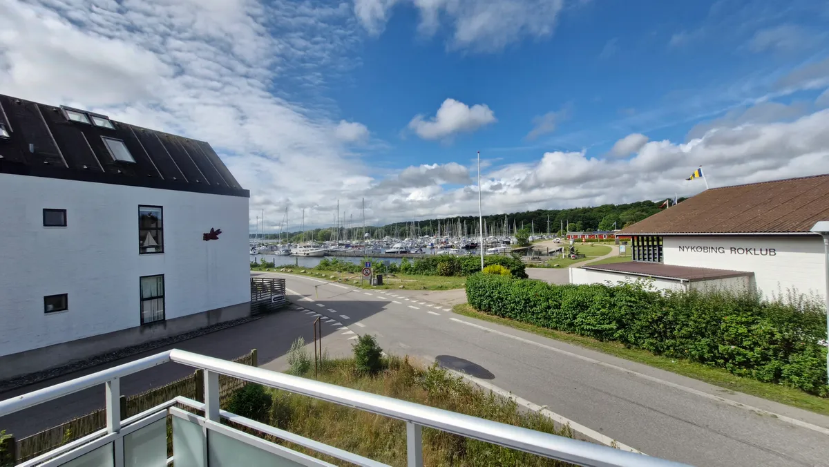 Et luftigt hav med adskillige både og Nykøbing Roklub på højre side af en asfaltvej med grønne buske og en bygning med rødt