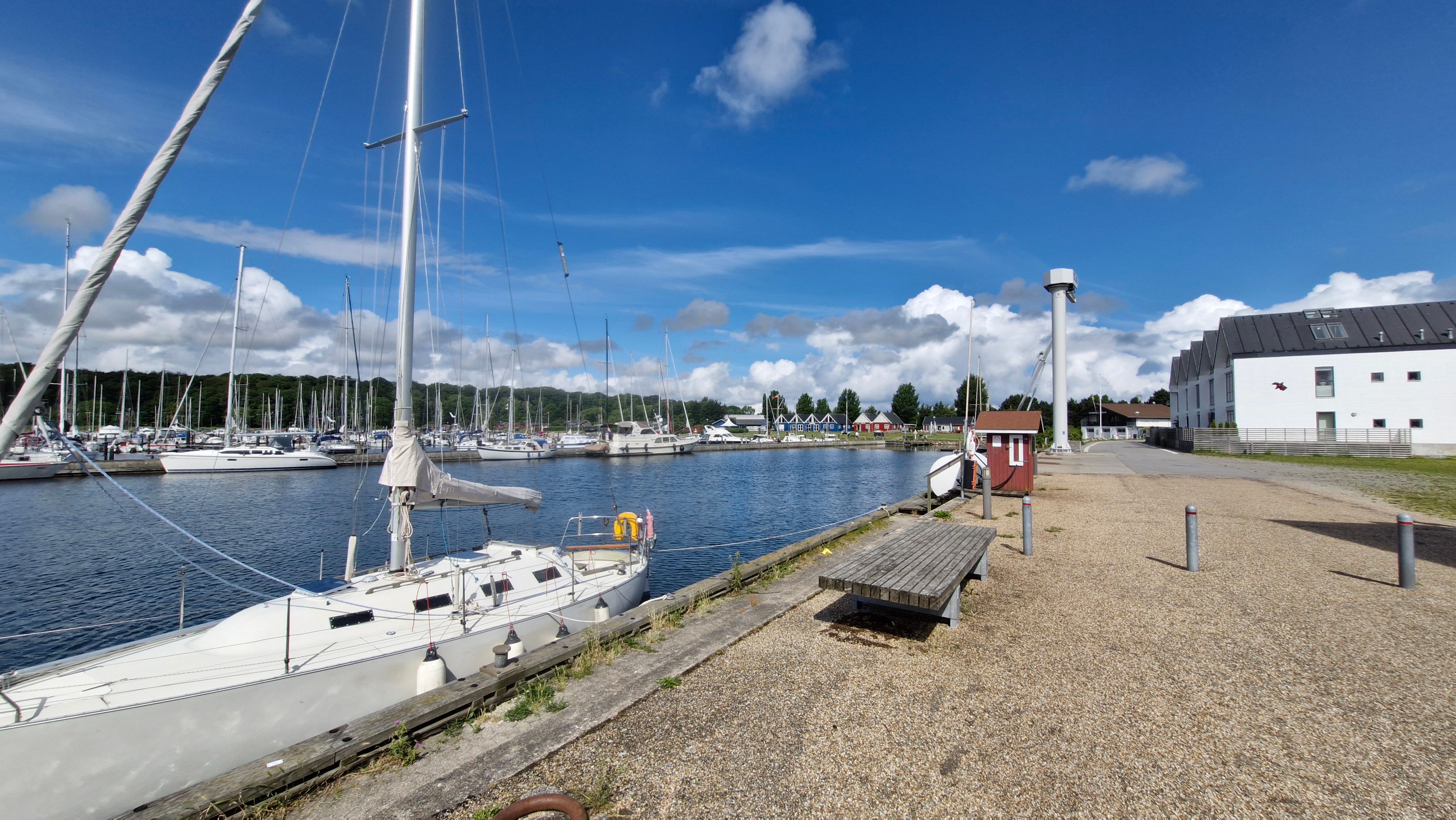 Et hvidt sejlskib er kædet til kajen i en havn med et gult redningsbøjle på skibet og andre skibe i baggrunden