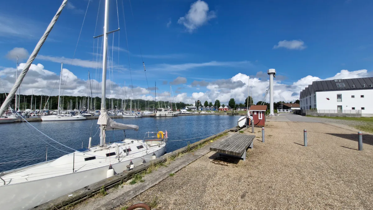 Et hvidt sejlskib er kædet til kajen i en havn med et gult redningsbøjle på skibet og andre skibe i baggrunden