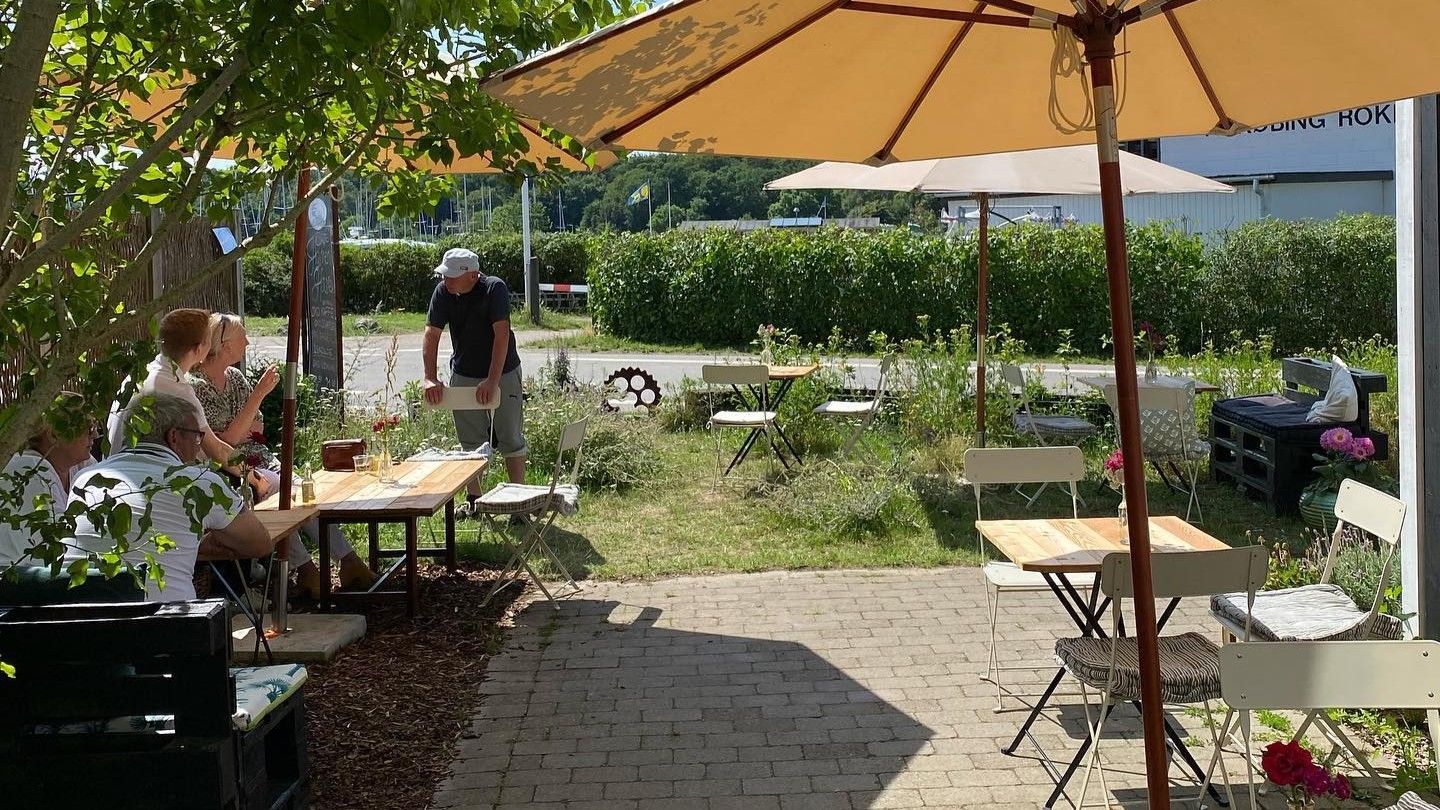 Mennesker sidder ved borde under parasoller på en grøn, blomstret gård. En mand står ved et bord med glas og et rødt