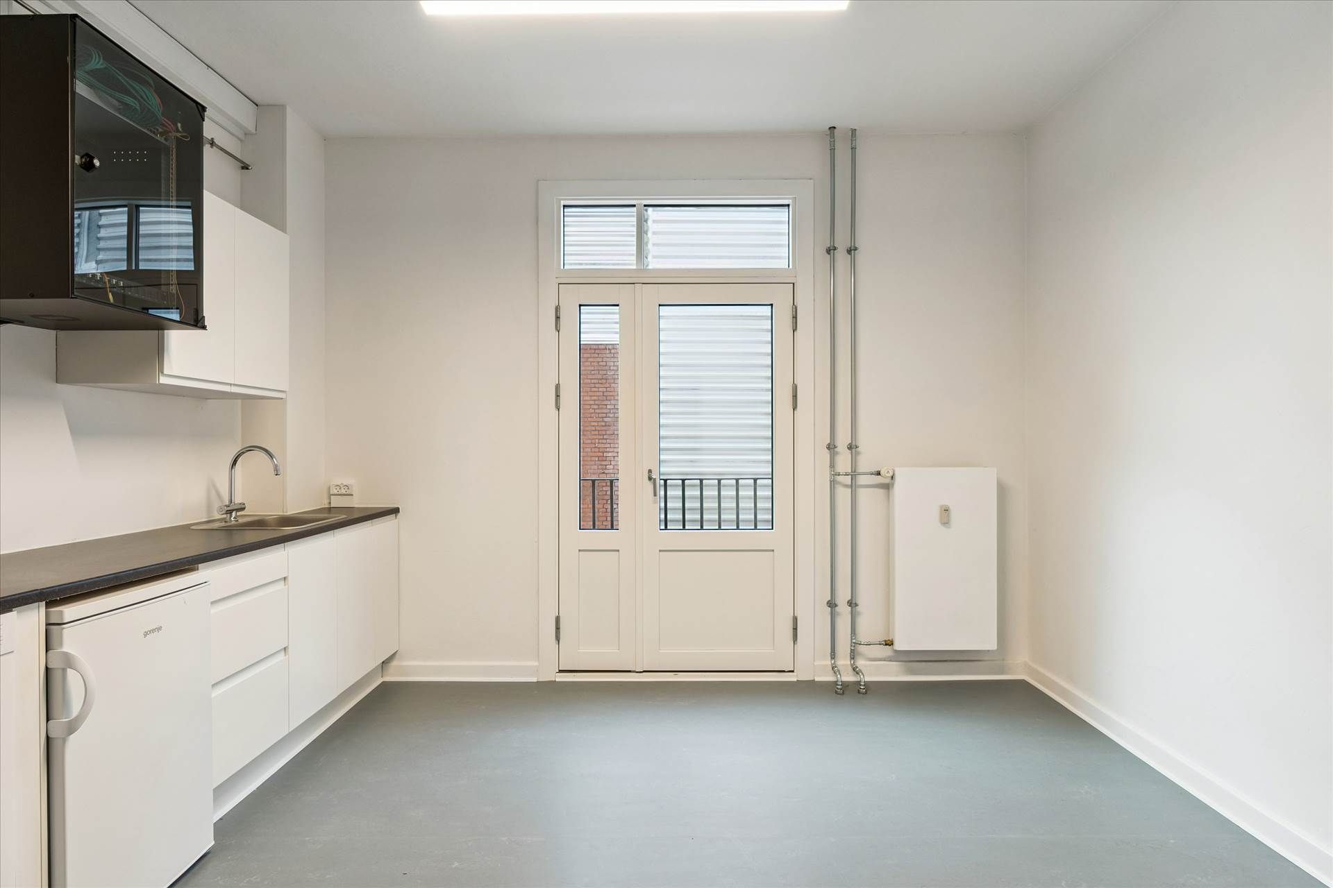 Et rent køkken med hvide vægge og møbler og gulv i grå og sort. Et hvidt køleskab og en sort hætte over ovnen. Et dobbeltdør