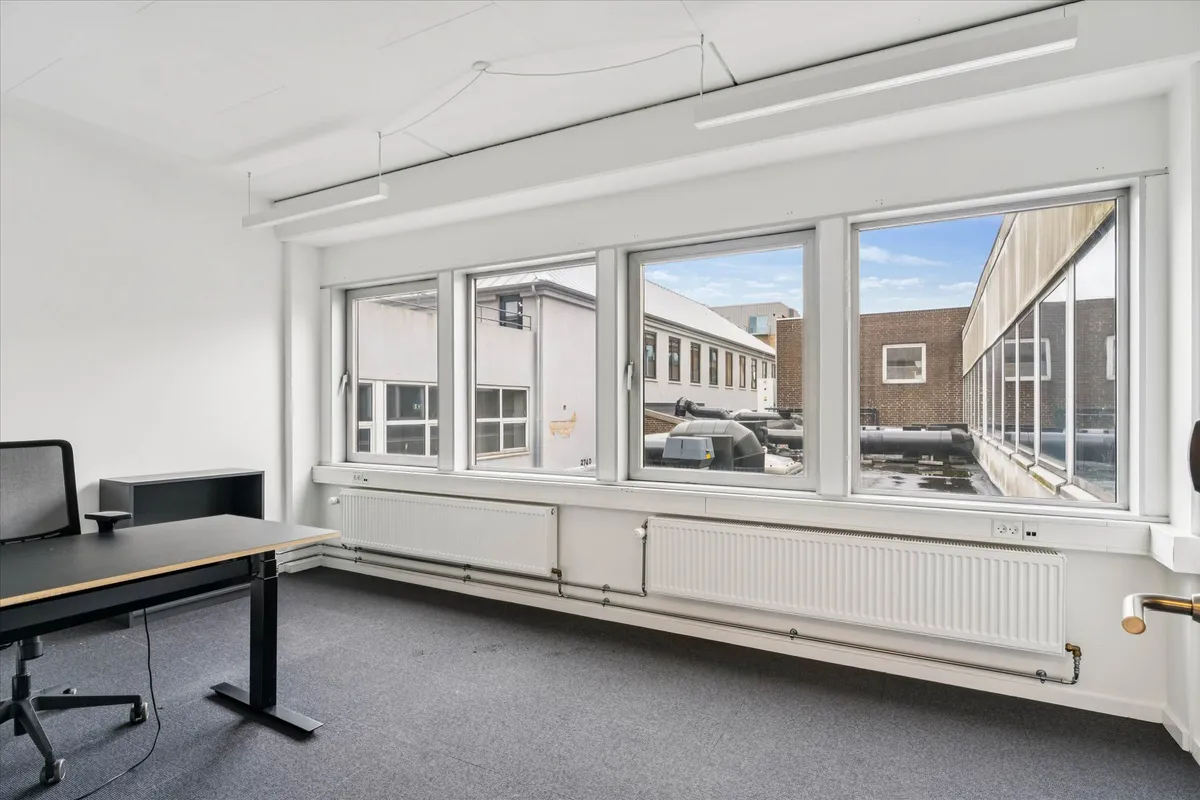 Værelse med skrivebord og kontormøbler samt radiatorer under vinduene og hvide vægge og loft