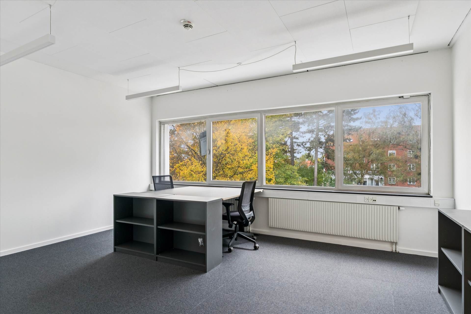 Kontorrum med høj synlighed, to skriveborde og vinduer med udsigt til bygninger og træer udenfor