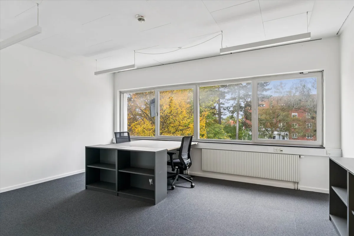 Kontorrum med høj synlighed, to skriveborde og vinduer med udsigt til bygninger og træer udenfor