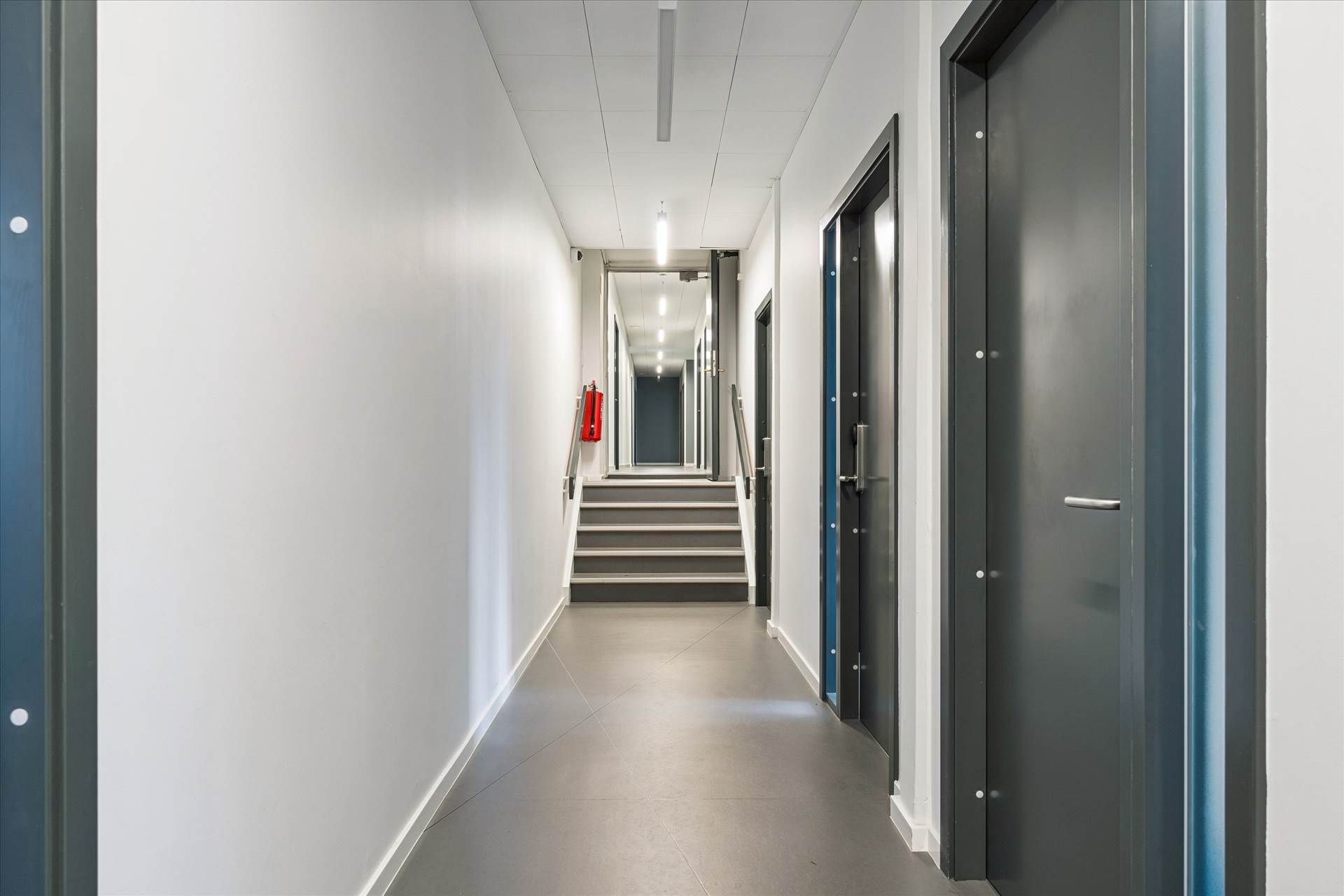 Hallen med lysegul gulvbelægning og hvide vægge og tre døre på højre side samt en trappe i midten