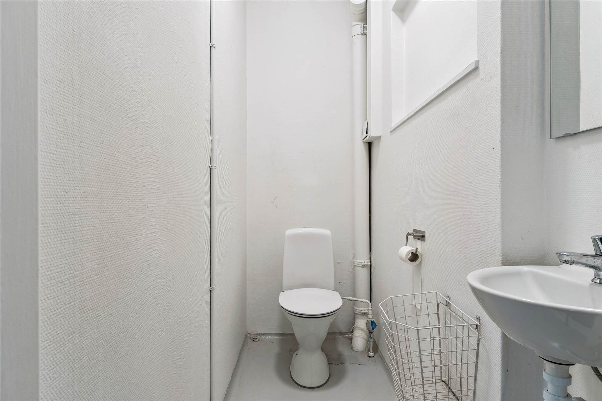 Værelse med toilet, hvidt badeværelse med toilet, hvidt håndvask og sæbe, samt en hvid papirhylde til håndklæder