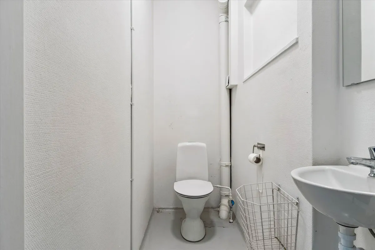 Værelse med toilet, hvidt badeværelse med toilet, hvidt håndvask og sæbe, samt en hvid papirhylde til håndklæder