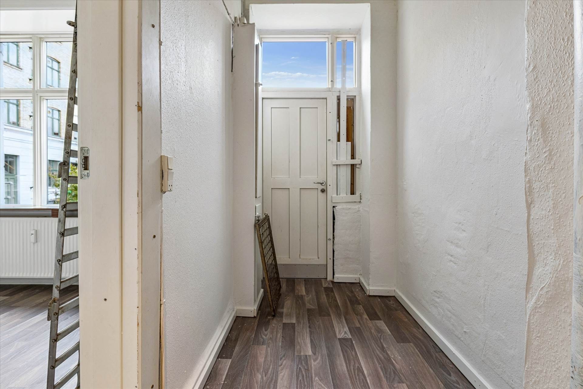 Korridor med hvide vægge, trægulv, hvid dør og lys fra vinduet, der er åben og vender mod en bygning med glasruder