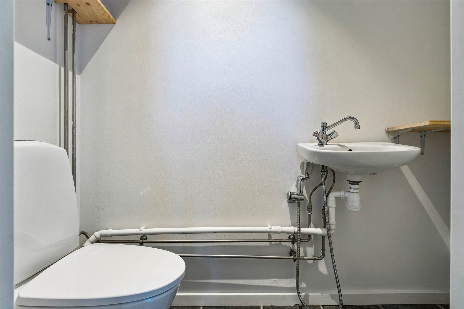 Lukket toilet med hvid toiletbøsse og hvid vask i et lille badeværelse med hvide vægge og gult gulv