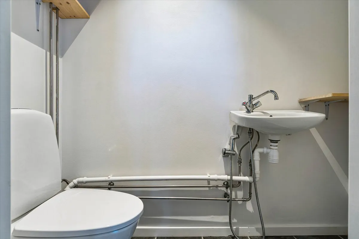 Lukket toilet med hvid toiletbøsse og hvid vask i et lille badeværelse med hvide vægge og gult gulv