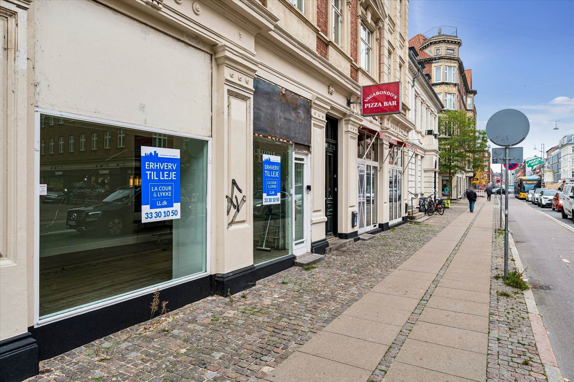 Butikslokal med udsigt til Amagerbrogade og tekst på facaden om til leje. På siden står Vagabondos Pizza Bar og et skilt med
