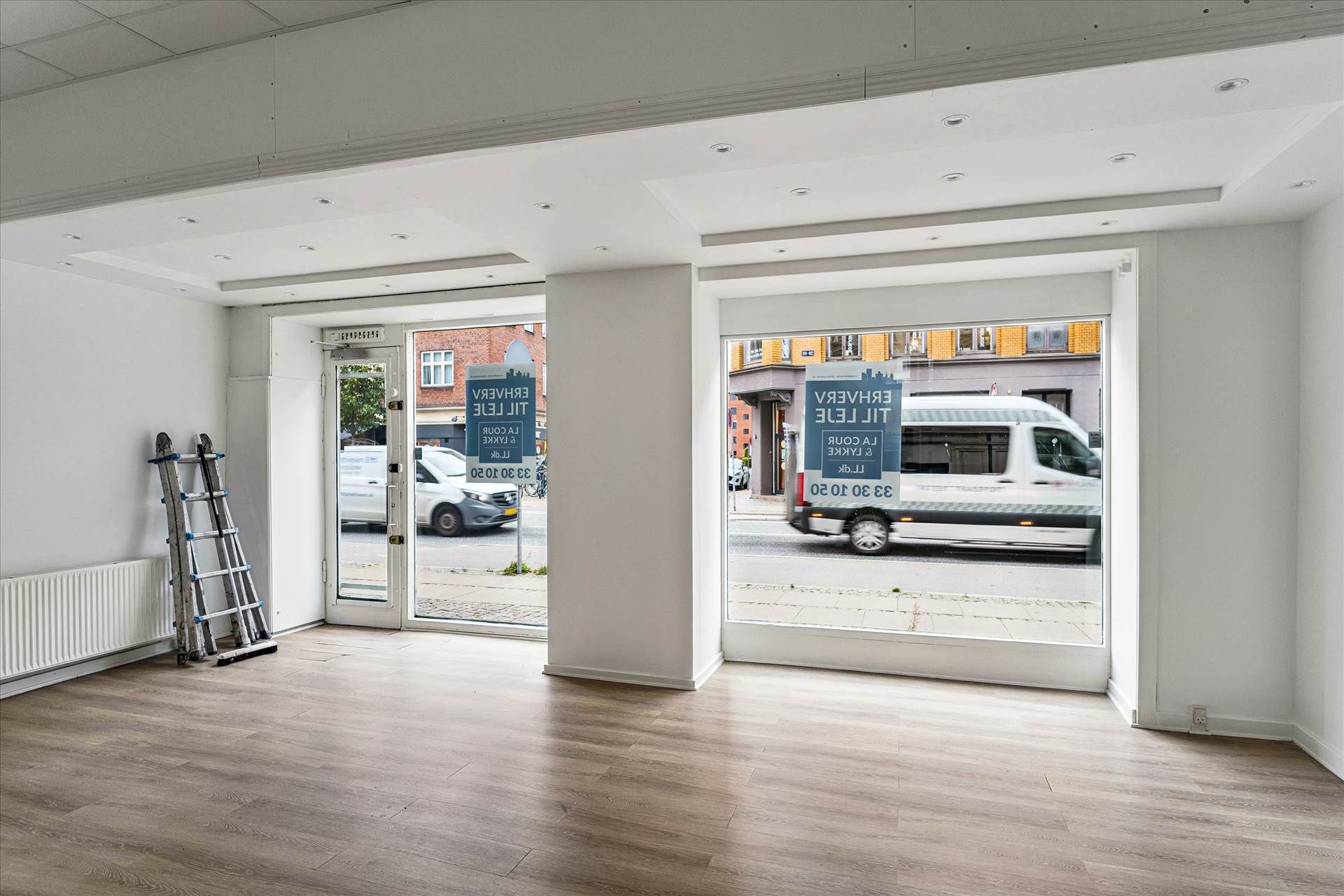 Butikslokal med store vinduer og trægulv. Vindueskiltet er blåt og hvidt. Vandkøler og stige er placeret mod væggen. To