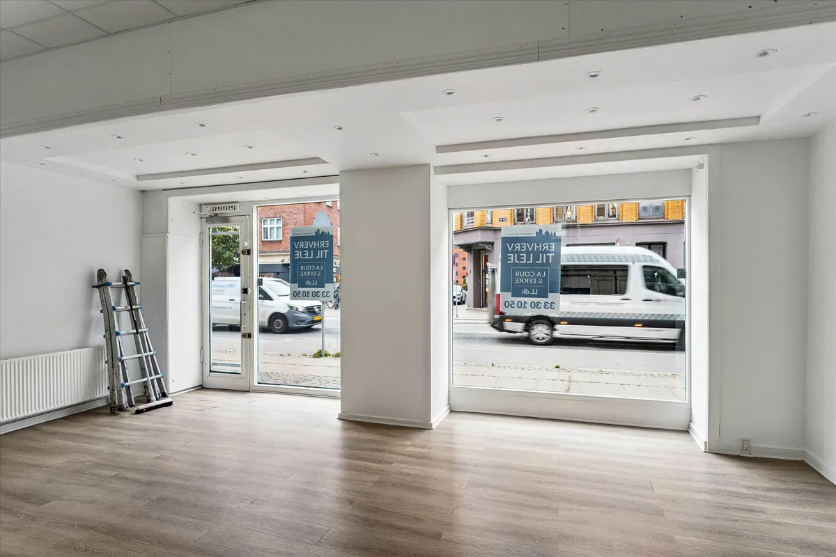 Butikslokal med store vinduer og trægulv. Vindueskiltet er blåt og hvidt. Vandkøler og stige er placeret mod væggen. To