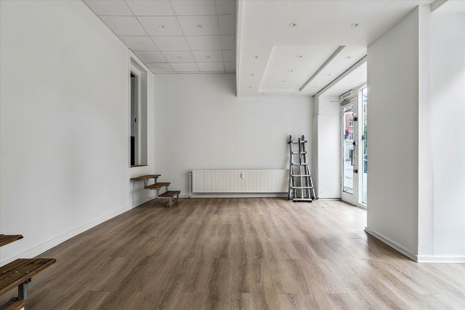 Et tomt showroom med højt til loftet og hårdt, trægulv i grå nuancer. Ståbord, trapper og en stige mod væggen